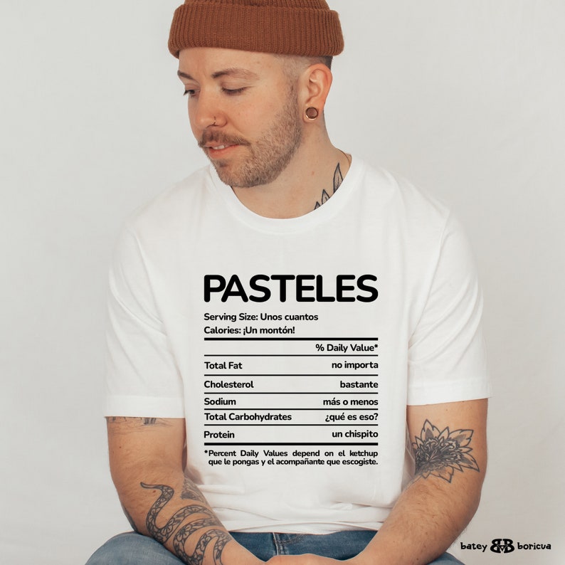 May include: A white t-shirt with a black graphic of a nutrition facts label for "PASTELES". The label says "Serving Size: Unos cuantos" and "Calories: &iexcl;Un mont&oacute;n!" with a disclaimer at the bottom that says "*Percent Daily Values depend on el ketchup que le pongas y el acompa&ntilde;ante que escogiste."