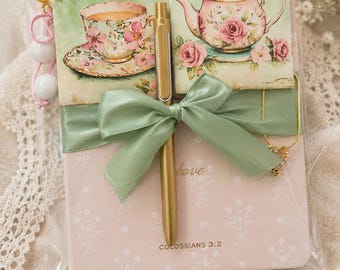 May include: A pink notebook with a floral tea cup and teapot illustration, wrapped in a sage green ribbon. A gold pen is tucked under the ribbon. The cover features the text "COLOSSIANS 3:2" and a small gold keychain with white beads attached to the side.