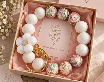 May include: A keychain featuring white silicone beads, floral patterned beads, and a white flower-shaped bead with a gold-toned metal ring. The keychain is presented in a light pink gift box with a card that reads Small Details Brighter Days.
