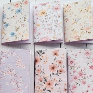 May include: A collection of small notebooks with floral patterns. The covers feature various pastel colors, including lavender, blue, and peach, adorned with delicate flower designs. The notebooks are open, revealing blank pages.
