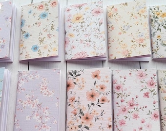 May include: A collection of small notebooks with floral patterns. The covers feature various pastel colors, including lavender, blue, and peach, adorned with delicate flower designs. The notebooks are open, revealing blank pages.