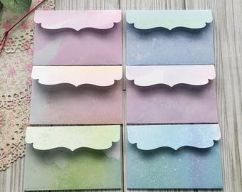 May include: A collection of pastel-colored envelopes with scalloped edges. The envelopes are in shades of pink, purple, blue, and green, with a watercolor effect. The envelopes are stacked and arranged in a grid pattern, suitable for stationery or small gifts.