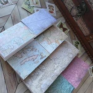 May include: A stack of vintage-style notebooks with map designs in various colors, including blue, beige, and pink. The notebooks are arranged on a wooden surface, with scattered postage stamps and a wooden chest in the background.