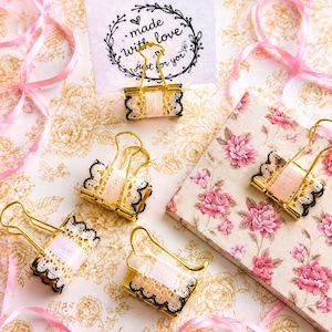 May include: Gold metal binder clips decorated with pale pink ribbon and black-edged white lace trim. One clip holds a white rectangular card featuring a black floral wreath design and the text "made with love just for you" in cursive script.