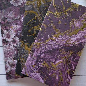 Puede incluir: Tres cuadernos decorativos con cubiertas únicas. Uno presenta un patrón floral en tonos morados y blancos. Los otros dos tienen diseños veteados en negro y morado con detalles dorados. Los cuadernos están dispuestos sobre una superficie de madera blanca.
