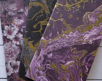 May include: Three decorative notebooks with unique covers. One features a floral pattern in shades of purple and white. The other two have marbled designs in black and purple with gold accents. The notebooks are arranged on a white wooden surface.