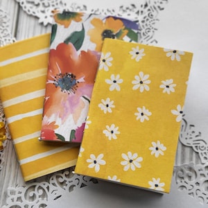 May include: Three small notebooks with floral and striped designs. One notebook has a yellow background with white daisies, another has orange and pink flowers, and the third has yellow stripes. The notebooks are arranged on a white lace doily.