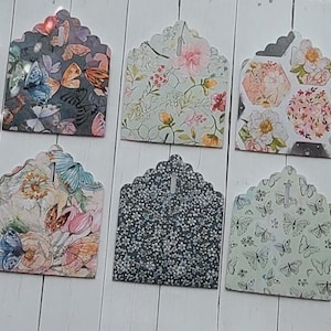 Può includere: Una collezione di dieci etichette decorative di carta con bordi smerlati. Ogni etichetta presenta un motivo floreale o a farfalla unico in vari colori, tra cui rosa, blu e verde. Le etichette sono disposte su una superficie di legno bianco.