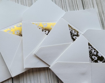 May include: A collection of white envelopes with a gold and floral patterned interior. The envelopes are arranged on a light gray wooden surface, showcasing their design and texture. The envelopes are open, revealing the decorative interior.
