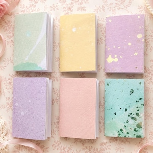 May include: Six small rectangular notebooks arranged in two rows of three. The covers feature various pastel colours including light green, yellow, lavender, light purple, soft pink, and teal, each with unique speckled or watercolour-style patterns.