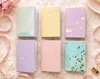 May include: Six small rectangular notebooks arranged in two rows of three. The covers feature various pastel colors including light green, yellow, lavender, light purple, soft pink, and teal, each with unique speckled or watercolor-style patterns.