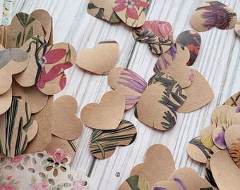 May include: A collection of heart-shaped paper cutouts with floral patterns in various colors, including red, purple, and yellow. Some hearts are plain, while others feature botanical designs. The cutouts are scattered on a light wooden surface.