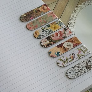 May include: A set of seven rectangular bookmarks with rounded ends, featuring various printed designs including floral patterns, wood grain, a small bird on a branch, and white lace motifs, arranged diagonally across a lined white notebook page.