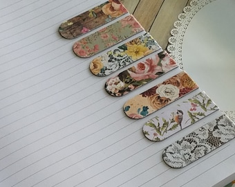 May include: A set of seven rectangular bookmarks with rounded ends, featuring various printed designs including floral patterns, wood grain, a small bird on a branch, and white lace motifs, arranged diagonally across a lined white notebook page.
