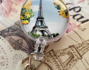May include: A circular badge reel featuring a photographic print of the Eiffel Tower in Paris, framed by yellow flowers and green foliage. The clear plastic housing includes a retractable cord, a metal swivel clip, and a clear vinyl strap with a metal snap.