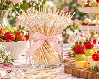 May include: A clear glass jar filled with wooden skewers topped with white faux pearls, tied with a light pink satin ribbon. The skewers are arranged upright in the jar, with additional skewers featuring pearls lying on a floral patterned plate.