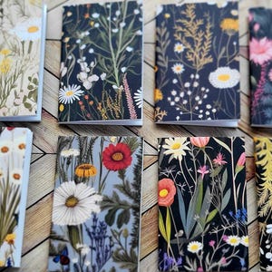May include: Eight small notebooks with floral designs. The covers feature various flower illustrations, including daisies, poppies, and other wildflowers, set against dark and light backgrounds. The notebooks are arranged on a wooden surface.