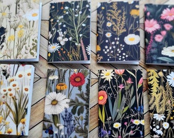 May include: Eight small notebooks with floral designs. The covers feature various flower illustrations, including daisies, poppies, and other wildflowers, set against dark and light backgrounds. The notebooks are arranged on a wooden surface.
