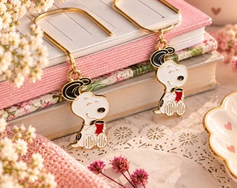 May include: A pair of gold-toned metal drop earrings featuring enamel charms of a white cartoon dog with black ears holding a small red mug. The earrings have a simple hook design and are displayed resting on top of a stack of closed notebooks.