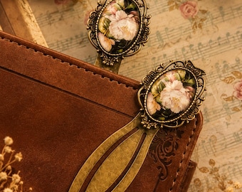 May include: Two antique-style brass bookmarks with floral oval inserts. The bookmarks are inserted into a brown leather journal. The background features sheet music and dried flowers, creating a vintage aesthetic.