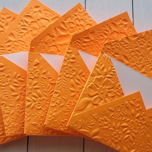 Puede incluir: Un conjunto de sobres de papel naranja en relieve con diseños florales. Los sobres están dispuestos en forma de abanico, revelando interiores blancos. El papel tiene una superficie texturizada, con hojas y flores detalladas.