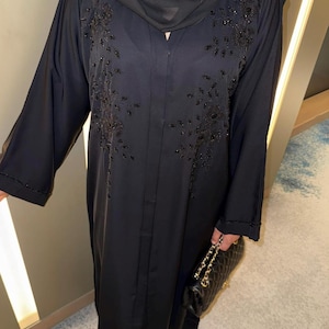 May include: A black abaya featuring long sleeves and intricate black beadwork embroidery on the chest area. The garment is paired with a matching black headscarf and a black quilted handbag with a gold chain strap, creating a monochromatic look.