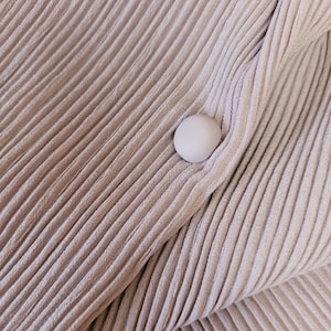 May include: A close-up of a light brown fabric with a ribbed texture. A single white button is sewn onto the fabric.