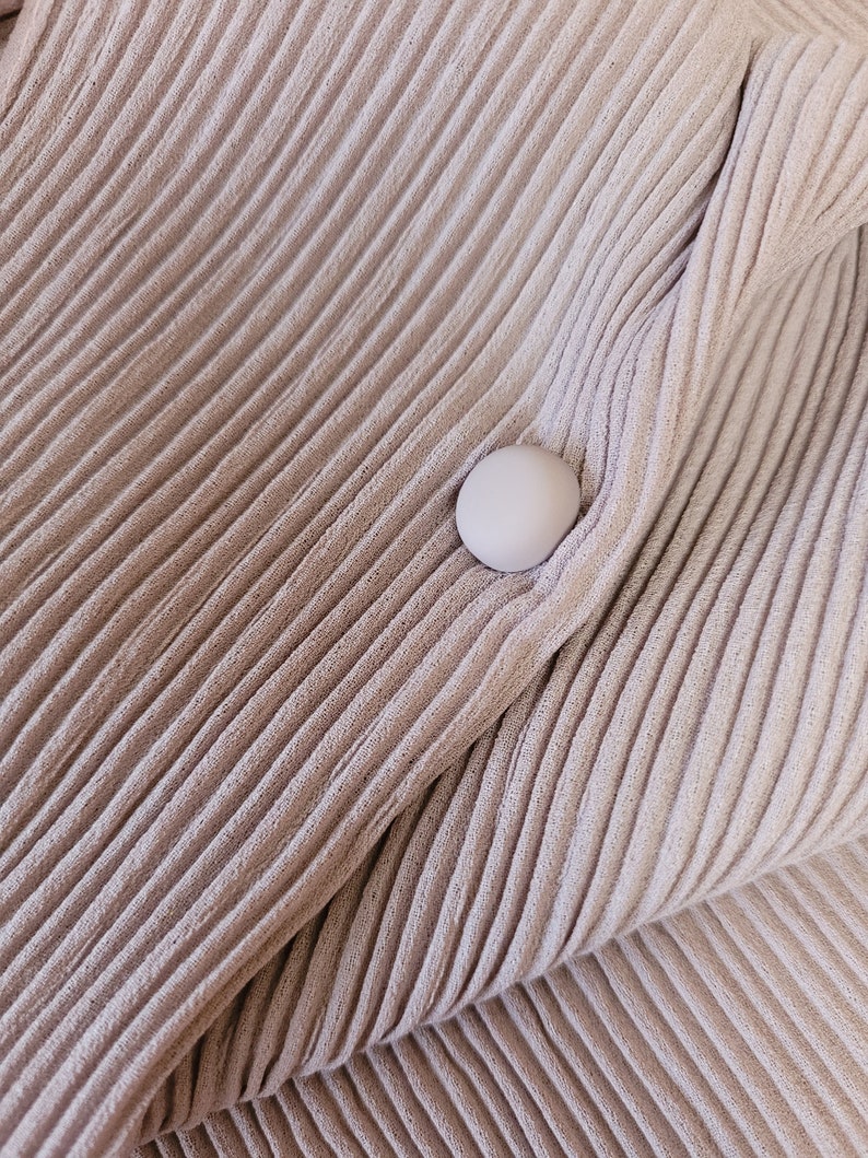 May include: A close-up of a light brown fabric with a ribbed texture. A single white button is sewn onto the fabric.