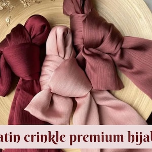 May include: Three satin crinkle premium hijabs in shades of burgundy and pink. The hijabs are arranged in a decorative knot on a wooden surface.