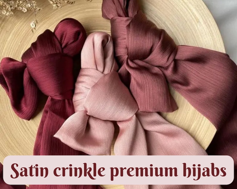 May include: Three satin crinkle premium hijabs in shades of burgundy and pink. The hijabs are arranged in a decorative knot on a wooden surface.