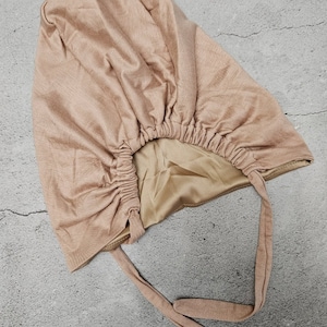 May include: A light brown satin-lined bonnet with a gathered elastic band and long ties. The bonnet is made of a textured fabric and is designed to protect hair while sleeping or during other activities.