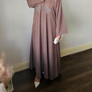 Puede incluir: Una abaya larga de color marrón con un diseño de manga larga. La abaya tiene un cuello en V y está decorada con bordados florales plateados. La abaya la lleva puesta una persona con zapatos blancos.