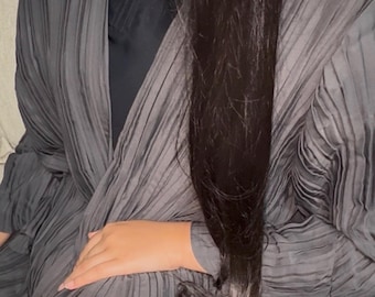 Könnte beinhalten: Ein dunkelgrauer, plissierter, vorne offener Mantel oder eine Abaya mit langen Ärmeln und einem strukturierten, vertikal gestreiften Stoffdesign. Das Kleidungsstück wird über einer einfarbigen schwarzen Basisschicht mit einem kleinen, runden Metallknopf am Ausschnitt getragen.