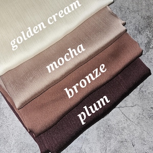 May include: Four fabric swatches stacked on top of each other. The swatches are in the colors golden cream, mocha, bronze, and plum.