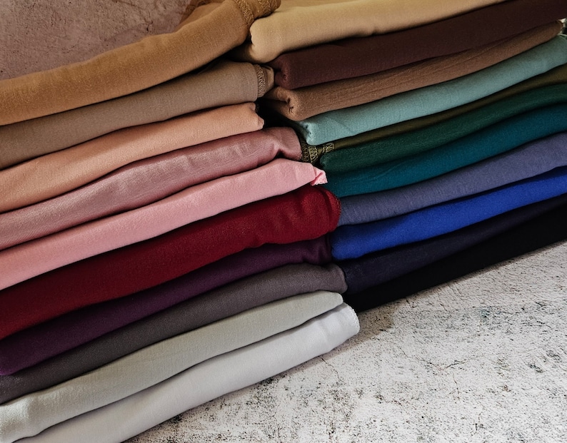 K&ouml;nnte beinhalten: Ein Stapel gefalteten Stoffes in verschiedenen Farben, darunter Beige, Rosa, Rot, Lila, Grau und Blau. Der Stoff ist in einem ordentlichen Stapel angeordnet, wobei die oberste Schicht &uuml;ber die unterste Schicht gefaltet ist.