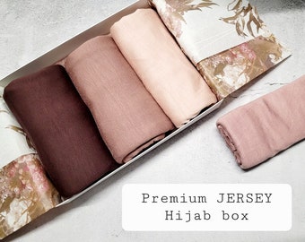 May include: A set of four hijab scarves in a white box with a floral pattern. The scarves are in shades of brown and pink. The box has the text "Premium JERSEY Hijab box" printed on it.