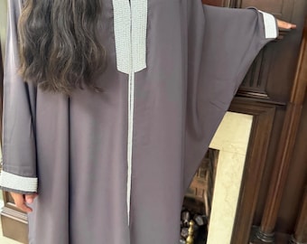 May include: A long, loose-fitting grey kaftan featuring wide sleeves and a prominent white beaded trim along the V-neckline and down the center front. The garment also includes matching white beaded detailing on the cuffs of the sleeves.
