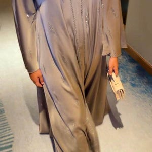 以下が含まれることがあります： 床まで届く長さの長袖トープカラーのカフタン。中央に縦方向の刺繍パネルが施され、生地全体にひし形のシルバーのスパンコールが散りばめられています。クリーム色の小さな構造的なクラッチバッグを合わせています。