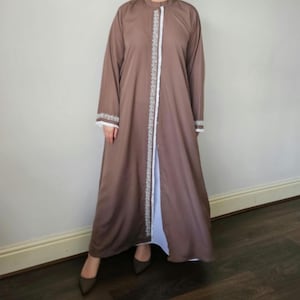 May include: A brown, long-sleeved abaya with white trim along the front and cuffs. The abaya is open in the front, revealing a white underlayer. The garment has a mandarin collar and is worn with brown heels.