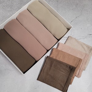 May include: A set of six rectangular scarves in various shades of brown and beige. The scarves are arranged in a white box and are folded neatly.