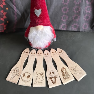 Peut inclure: Un gnome rouge avec une barbe blanche et un cœur gris sur son chapeau est placé au-dessus de sept spatules en bois. Chaque spatule présente un personnage différent d'un film d'animation populaire, gravé dans le bois.