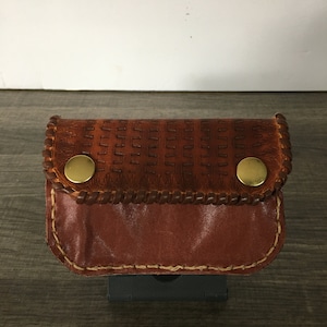 May include: A small brown leather pouch with two gold metal snaps. The pouch has a decorative stitched border and a textured surface.