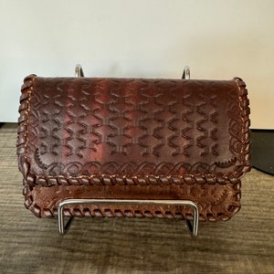 May include: A brown leather wallet with a geometric pattern and a braided trim. The wallet is on a metal stand.