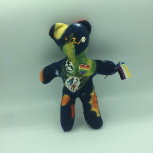 Peut inclure: Un ours en peluche avec un tissu à motifs bleus, verts et oranges. L'ours a un cœur arc-en-ciel, un bouton avec un symbole de paix et un petit drapeau avec un arc-en-ciel et des rayures noires et blanches cousus dessus.