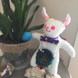 May include: White felt bunny with pink ears, holding a green and blue felt basket. The bunny has yellow and pink embroidered eyes and a purple and yellow polka dot bow tie. The bunny is standing on two gray rocks.