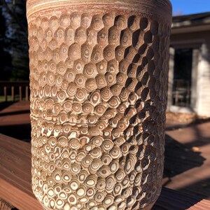 May include: A ceramic vase with a textured, honeycomb-like pattern in shades of brown and white. The vase has a cylindrical shape and a slightly flared rim.