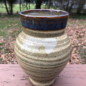 May include: A handmade ceramic vase with a wide, striped body and a flared rim. The vase features a color palette of tan, brown, and blue, with a speckled texture. The vase is approximately 8 inches tall.