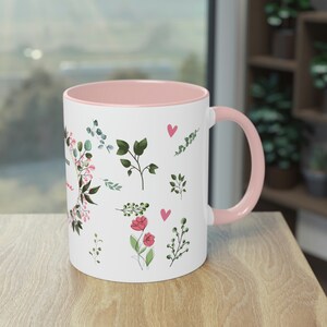 Può includere: Tazza in ceramica bianca con bordo e manico rosa. La tazza presenta un design floreale con fiori rosa, foglie verdi e cuori.