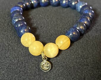 May include: A beaded bracelet featuring alternating dark blue and yellow spherical beads. A small silver charm with a crescent moon and star design hangs from the yellow beads. The bracelet is displayed on a dark gray surface.