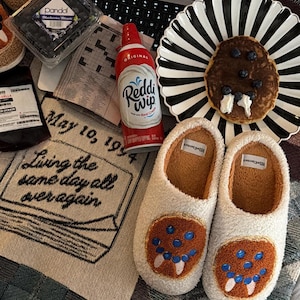 May include: A pair of white fuzzy slippers with a brown pancake graphic featuring blue berries and white cream. Nearby are a black and white striped plate with a pancake, a can of Reddi-wip, a container of blueberries, and a cloth with the text May 10, 1994 Living the same day all over again.